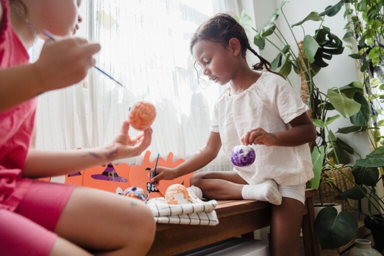 Child-Safe Halloween Decorations: 5 Essential CPSC Rules for Low-Risk Holiday Styling 2 Two children painting small pumpkins orange and purple as a Halloween craft activity by a sunny window